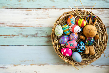 flat lay of colorful vibrant easter eggs with stripe flower pattern paint, laying on basket nest with brown bunny rabbit soft toy, blue wooden table background representing celebrating easter holidays