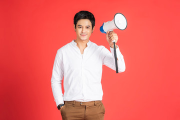 handsome asian businessman smiling holding megaphone to amplify sound, shouting announcing messages to alert people, hand in pocket wearing casual clothing, standing posing in red isolated background