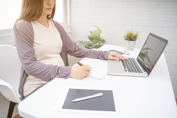 pregnant asian woman working from home in modern white office using pen writing on notepad with tablet laptop computer technology smiling with happiness of motherhood pregnancy expectant new born baby