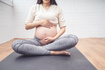 Body shot of pregnant woman caressing her baby belly, sitting on black yoga mat training yoga...