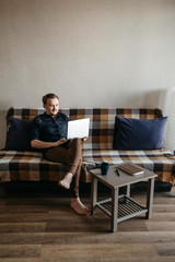 A young European guy in a dark blue shirt and brown trousers with a dark beard looks at a laptop. Sits on the couch, laptop on his knees. Home insulation. Home Office.