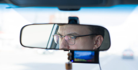 a young man with glasses at the wheel of a car is reflected in the rearview mirror