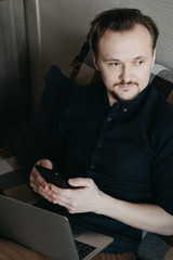 A young European man in a dark blue shirt and brown trousers with a dark beard is holding a telephone. Sits on the couch, laptop on his knees. Home insulation. Home Office.