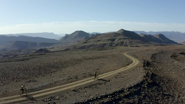 A low aerial flyover tracking two cyclists descending a barren desert ridge, bikepacking through a vast expanse of arid sand and rock and stark mountain ridges stretching as far as the eye can see.