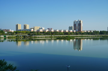 Fototapeta na wymiar Cityscape by the river under blue sky, buildings reflecting on peaceful water
