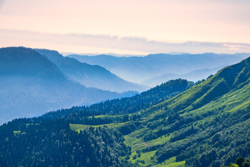 Obraz premium View over the Green Valley, surrounded by high mountains on a pink summer sunset. Layers of mountains in the haze during sunset.