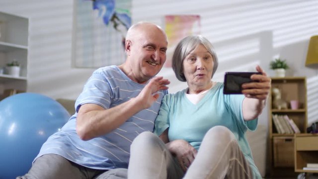 Happy senior couple in sportswear sitting on the floor in the living room, smiling and waving while taking a selfie with smartphone after home workout
