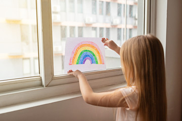 Kid painting rainbow during Covid-19 quarantine at home. Girl near window. Stay at home Social media campaign for coronavirus prevention, let's all be well, hope during coronavirus pandemic concept
