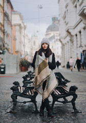 Fototapeta na wymiar A young caucasian girl in coat, angora hat, wide scarf is standing alone leaning on a cast-iron bench awaiting a date. Lviv, Ukraine.