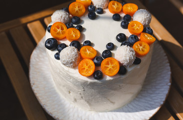 A birthday summer homemade cake with whipped curd cream frosting and decorated with kumquat, blueberries and coconut sweets. Served on white handmade plate on a light wooden table. Natural lighting