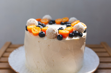 A summer homemade cake with white whipped curd cream and decorated with kumquat, blueberries and coconut sweets. Served on white plate on a light wooden table. Gray background. Natural lighting