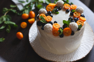 A celebratory fruit cake with whipped curd cream icing decorated with cut kumquats, blueberries, coconut sweets and mint leaves on a dark background. Selective focus