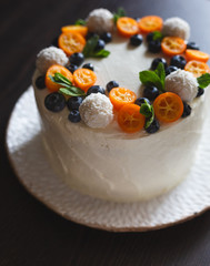 Summer homemade biscuit cake covered with whipped curd cream, decorated with fresh berries and fruits of kumquats, blueberries, coconut sweets and mint leaves on a white handmade plate