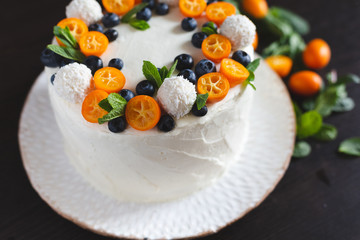 Cut view of a wedding or birthday summer cake covered with white whipped curd cream and decorated with kumquat, blueberries, coconut sweets and fresh mint leaves on a white plate on dark background