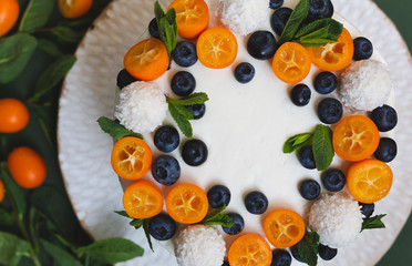 Cut view of a homemade birthday or wedding cake with cream cheese frosting on big white plate. Decorated with fresh blueberries, kumquats and mint leaves on a green background. Top view