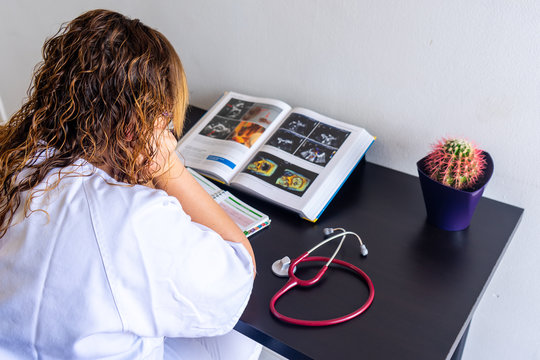 Young woman studying medicine sitting at a desk. There is also a cardiology textbook where you can see electrocardiograms.  On the desk there is a stethoscope and a cactus. 