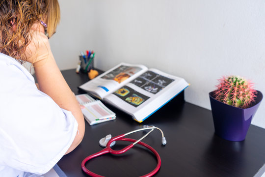 Young woman studying medicine sitting at a desk. There is also a cardiology textbook where you can see electrocardiograms.  On the desk there is a stethoscope and a cactus. 