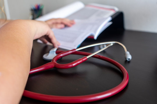 On a desk in the foreground a stethoscope. In the background is a pencil with pens, a cactus and a laptop. All out of focus.