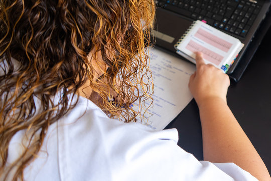 Young woman studying medicine sitting at a desk. On the desk is a laptop with an EKG on the screen. There is also a cardiology textbook where you can see electrocardiograms.  