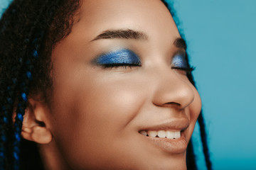 Cheerful afro american young woman smiling with closed eyes. Cut view of blue with metal color eyeshadows. Close up. Isolated over blue background.