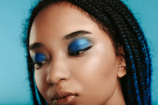 Close up of african model with stylish metal blue eyeshadows makeup look down. Calm peaceful fashionable model. Dreadlocks with blue tape in hair. Isolated over blue background.