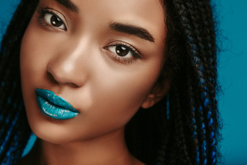 Calm peaceful attractive young afro american woman with blue lips posing on camera. Nude eyeshadows in stylish makeup. Isolated over blue background. - obrazy, fototapety, plakaty