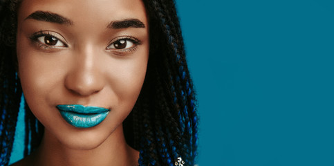 Cut view portrait of cheerful confident afro american woman smiling and looking on camera. Stylish girl with blup lipstick on lips and nude eyeshadows. Modern makeup. Isolated over blue background.