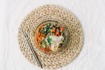 Japanese ramen soup in a glass plate with vegan ingredients, fresh vegetables in small plates, rice noodles, mushrooms and miso paste broth, on a natural wicker napkin on a white table