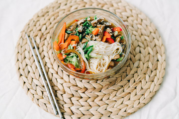 Obraz premium Japanese ramen soup in a glass plate with vegan ingredients, fresh vegetables in small plates, rice noodles, mushrooms and miso paste broth, on a natural wicker napkin on a white table