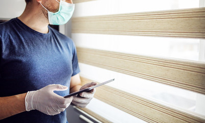 Young man with mask and gloves typing on tablet, searching for news online. Be responsible, stay at...