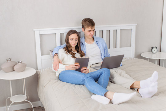 Young Couple Using On Laptop Computer And Ipad, Resting Together On Day And Weekend Off At Home Bed. Serious, Not Talking, Little Communication Because Of Gadgets. Man And Woman Choose Film, Goods, Gi