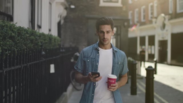 Handsome young man walking the city using mobile phone. Attractive guy using a smartphone and drinking a cup of takeaway coffee. Slow motion.