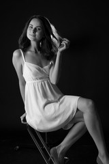 stunning black and white portrait of a sexy young brunette girl in a white midi dress sitting on a high chair on a black background.  Low kay. Beauty and Fashion