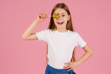 Portrait of a surprised little girl holding heart shaped lollipop and looking at camera isolated over pink background.
