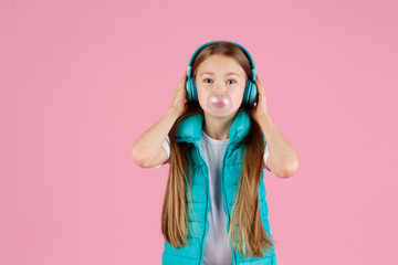 A little girl with headphones explodes pink chewing gum on a pink background