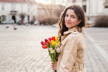 Beautiful young woman with spring tulips flowers bouquet at city street. Happy girl walking outdoors. Spring portrait of pretty female in old town