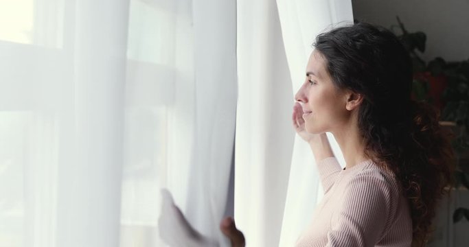 Happy optimistic young woman opening curtain lace looking through window. Smiling confident lady enjoying watching beautiful cityscape view, feeling hope and dreaming of good future at home.