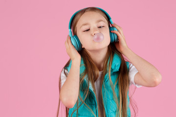 A little girl with headphones explodes pink chewing gum on a pink background