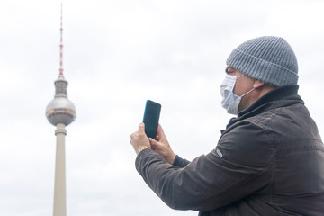 Disease outbreak, coronavirus covid-19 pandemic. Portrait of adult man wearing medical protective mask on his face taking a picture of the Berlin Television Tower with his smartphone