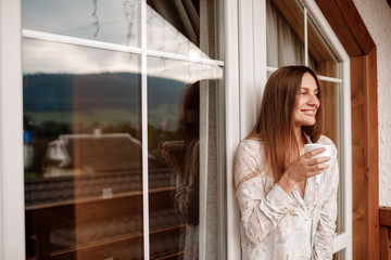 portrait of young woman on the balcony holding a cup of coffee or tea in the morning. She in hotel room looking at the nature in summer. Girl is dressed in stylish nightwear. Relax time