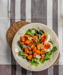 Roasted veggie salad. Oven baked butternut squash, lettuce, boiled egg and mixed seeds salad on a vintage white plate served on top of a wooden table. Healthy and colourful salad. Top view. 