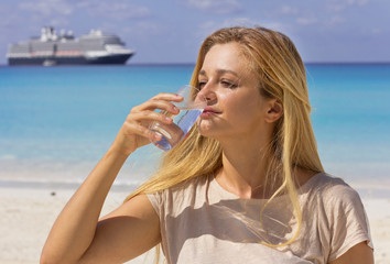 Young beautiful girl drinks water on the beach. Woman with a glass of water. Hot beach day.