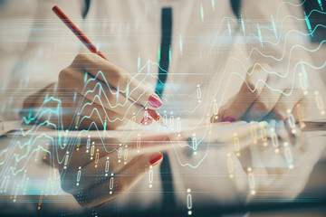 A woman hands writing information about stock market in notepad. Forex chart holograms in front. Concept of research.