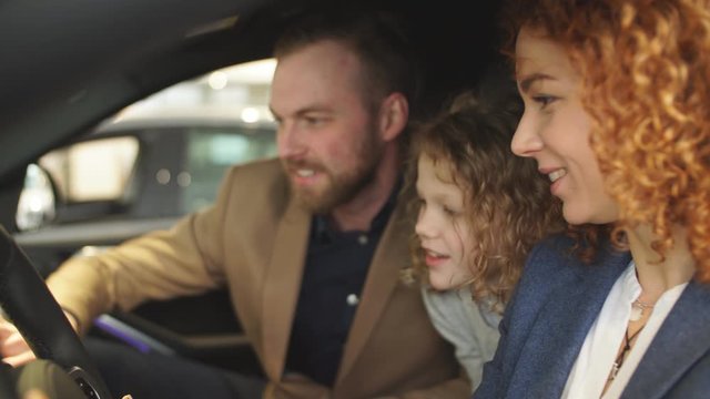 Young beautiful caucasian family try new car from inside. Man, woman and child sit inside of car and examine its features. Child want parents to buy it.