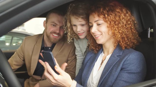 Young beautiful caucasian family try new car from inside. Man, woman and child sit inside of car and examine its features. Child want parents to buy it.