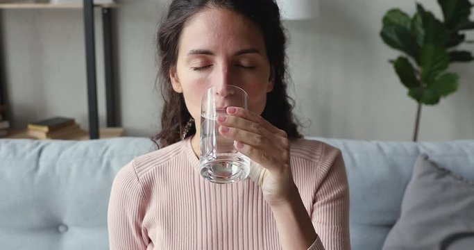 Thirsty young woman holding glass drinking fresh mineral water to stay healthy at home sitting on sofa. Dehydrated ill lady hydrating thirst keeping fluid balance, illness recovery, healthcare concept