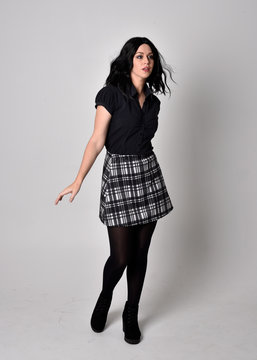 Portrait of a goth girl with dark hair wearing blue and plaid skirt with boots. Full length standing pose on a studio background.
