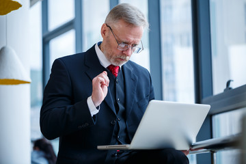 Smiling happy managing director thinks about his successful career development while standing with a laptop in his office near the background of a window with copy space