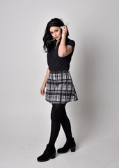 Portrait of a goth girl with dark hair wearing blue and plaid skirt with boots. Full length standing pose on a studio background.