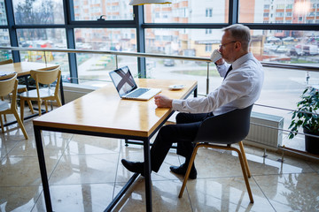 Male freelancer is working in a cafe on a new business project. Sits at a large window at the table. Looks at a laptop screen with a cup of coffee